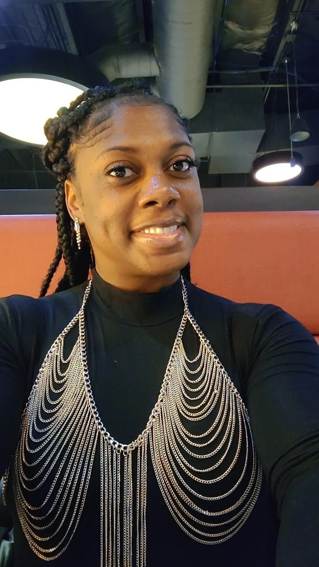 Woman smiling, wearing a black top with layered chain necklace in a brightly lit room.